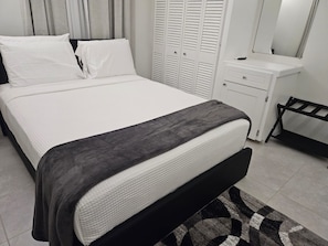 Una cama bien hecha con colchón blanco y manta gris, una mesita de noche blanca con espejo, y un banco negro.