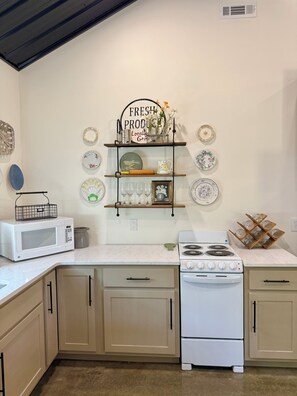 A kitchen with a white stove, a microwave, and a sink. There are shelves with plates and a sign that says 'FRESH PRODUCE'.