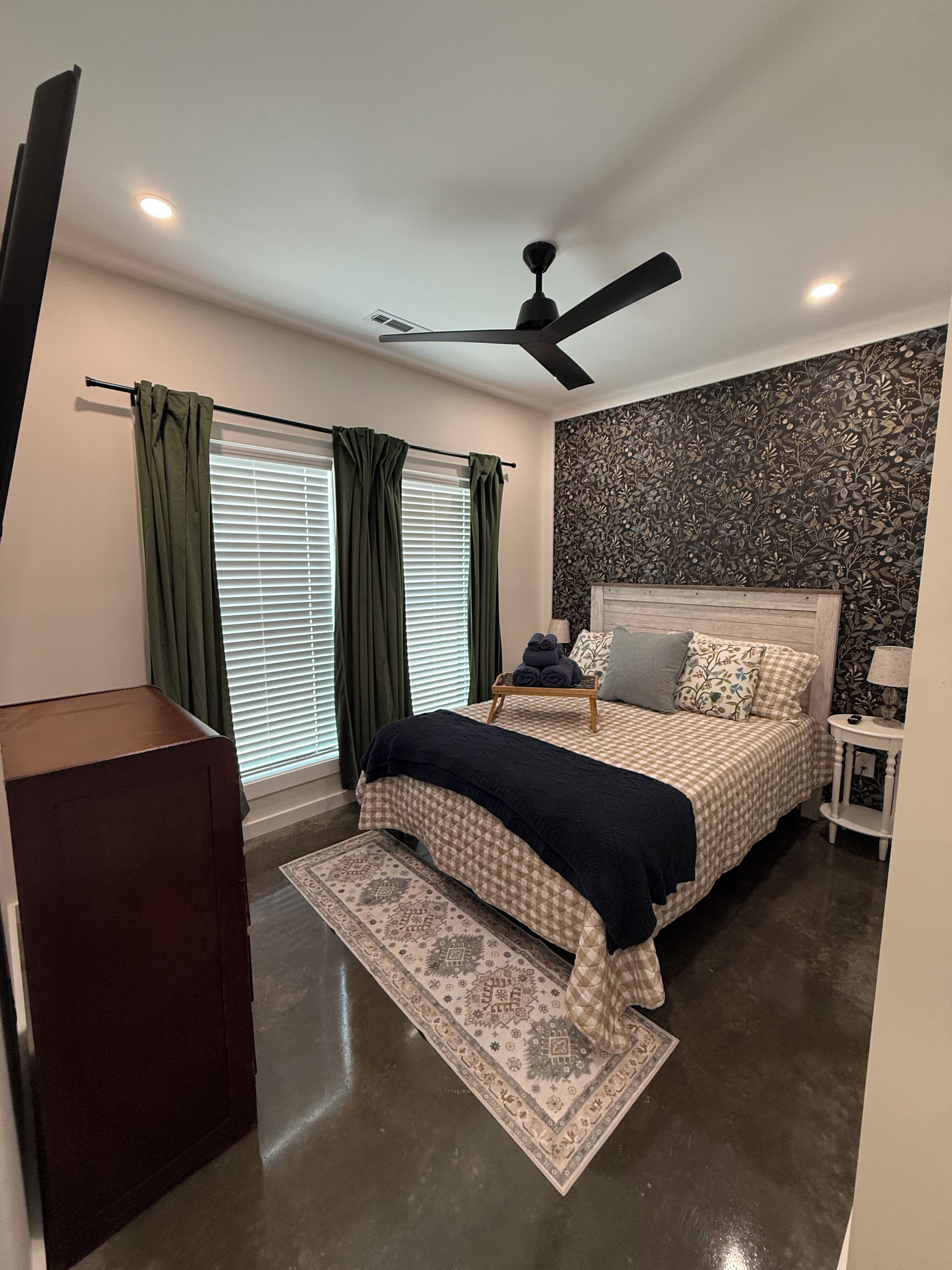A bedroom with a bed, a ceiling fan, a window with curtains, a wall with a floral pattern, and a small bedside table with a lamp.