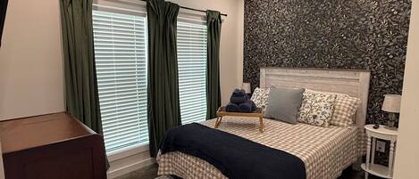 A bedroom with a bed, a ceiling fan, a window with curtains, a wall with a floral pattern, and a small bedside table with a lamp.