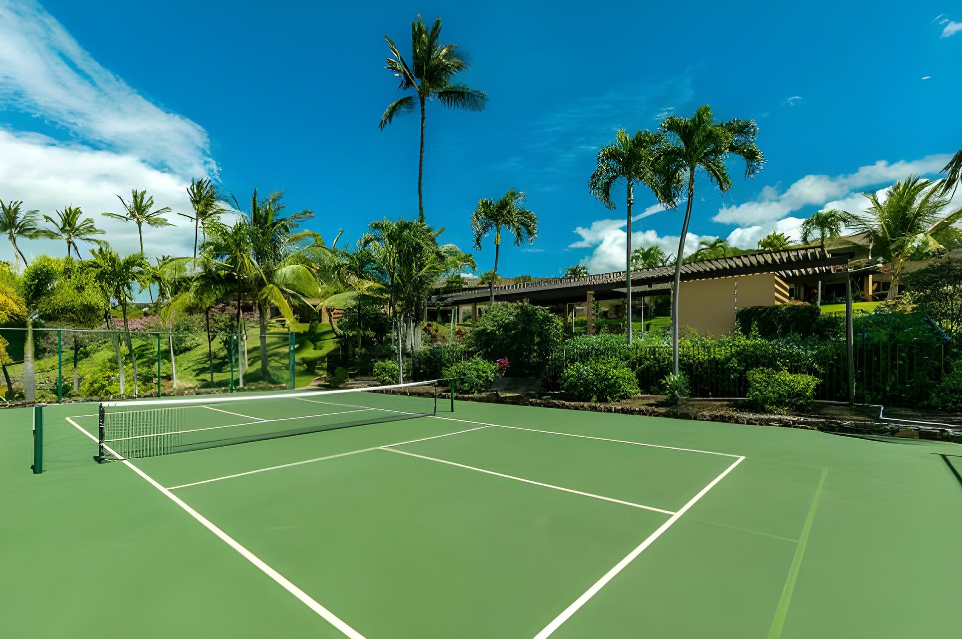 A tennis court with a net, surrounded by palm trees and a building with a covered walkway.