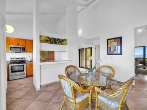 A modern kitchen with a microwave, oven, and wooden cabinets. There is a dining area with a glass table and wicker chairs. - Wailea Ekolu | 4BR | Sleeps 12 (Kihei)
