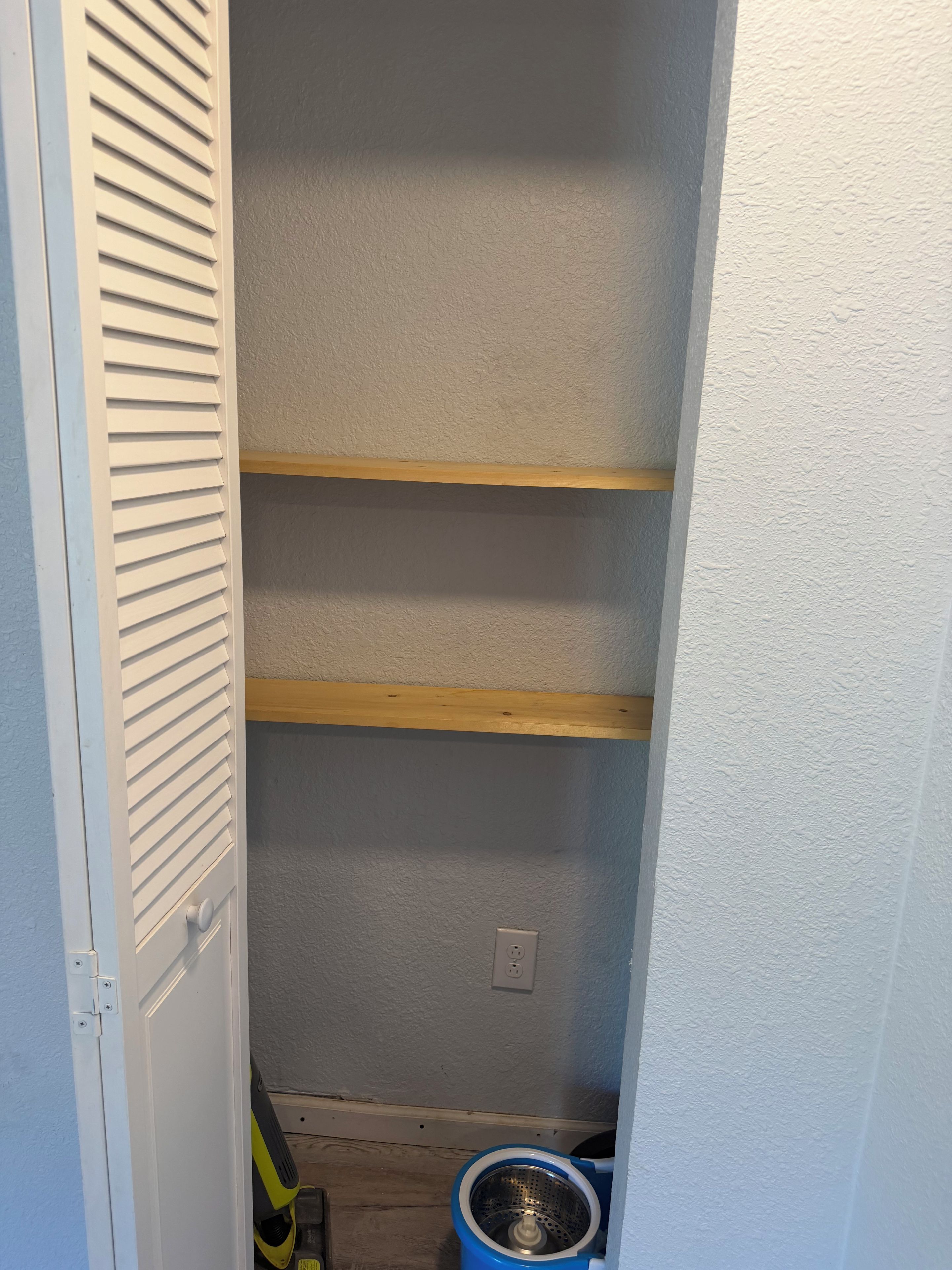 A narrow hallway with a white door on the left, a blue trash bin, and a wooden shelf unit against the wall.