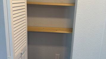 A narrow hallway with a white door on the left, a blue trash bin, and a wooden shelf unit against the wall.