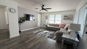 A modern living room with a sectional sofa, a flat-screen TV, a ceiling fan, and a decorative rug. - Beautiful 2-bedroom house with AC in charming New Port Richey (New Port Richey)