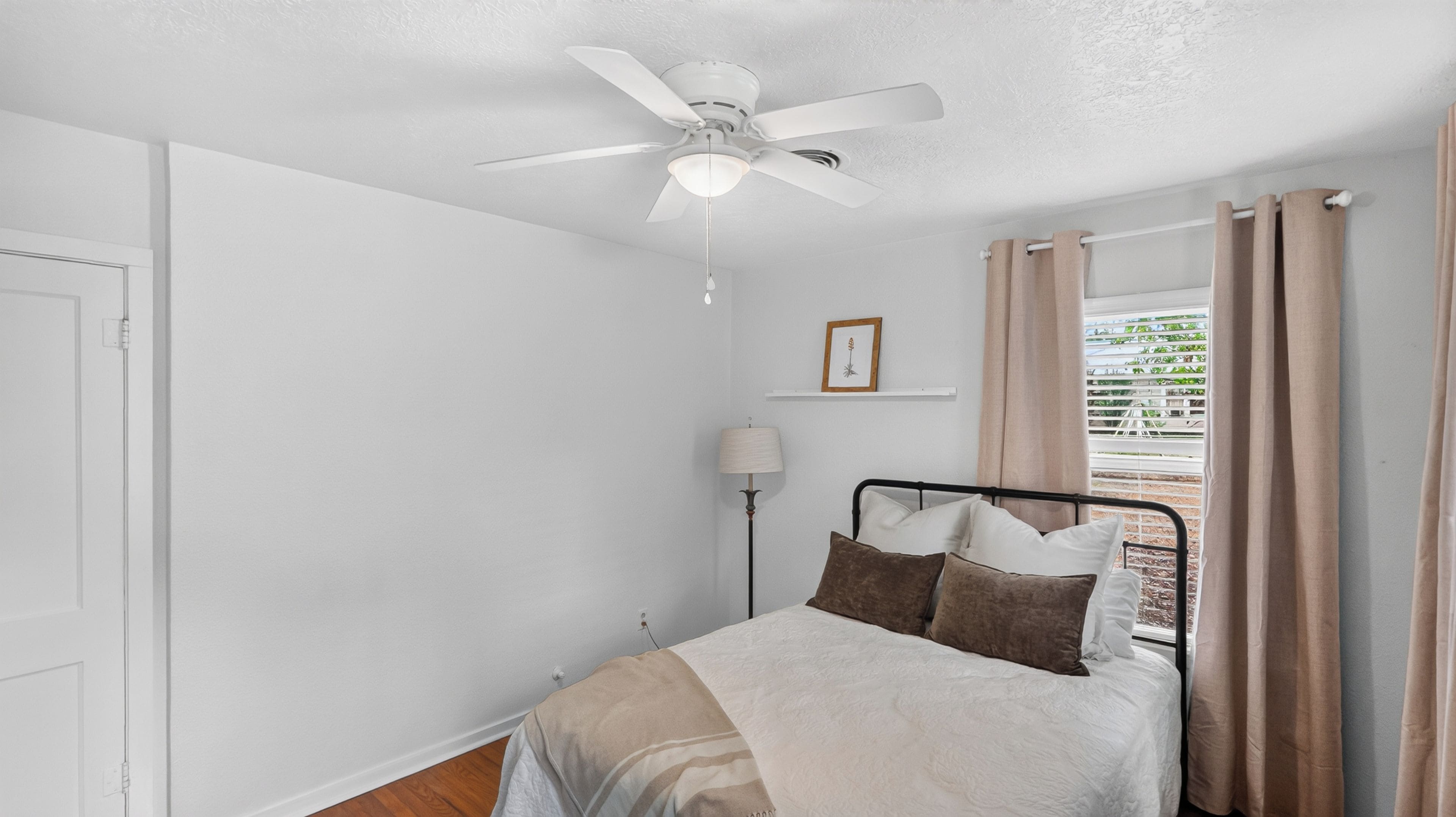 Une chambre à coucher avec un lit, un ventilateur de plafond, une lampe de chevet, une fenêtre avec des rideaux et un tableau encadré au mur.