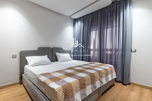 Quarto moderno com cama, cabeceira cinza, travesseiros brancos, manta xadrez e janela com cortinas cinzas.