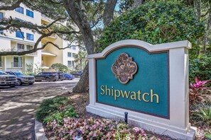 A sign with the name 'Shipwatch' in gold lettering on a teal background, surrounded by greenery and a parking area with cars.