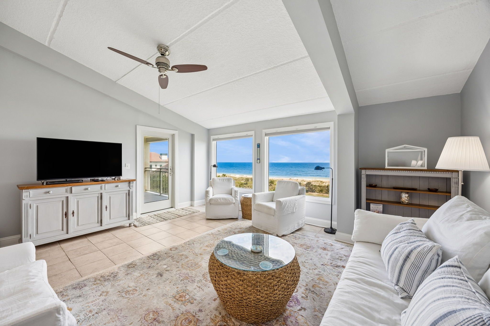 A spacious living room with a television, white furniture, a ceiling fan, and a view of the ocean.