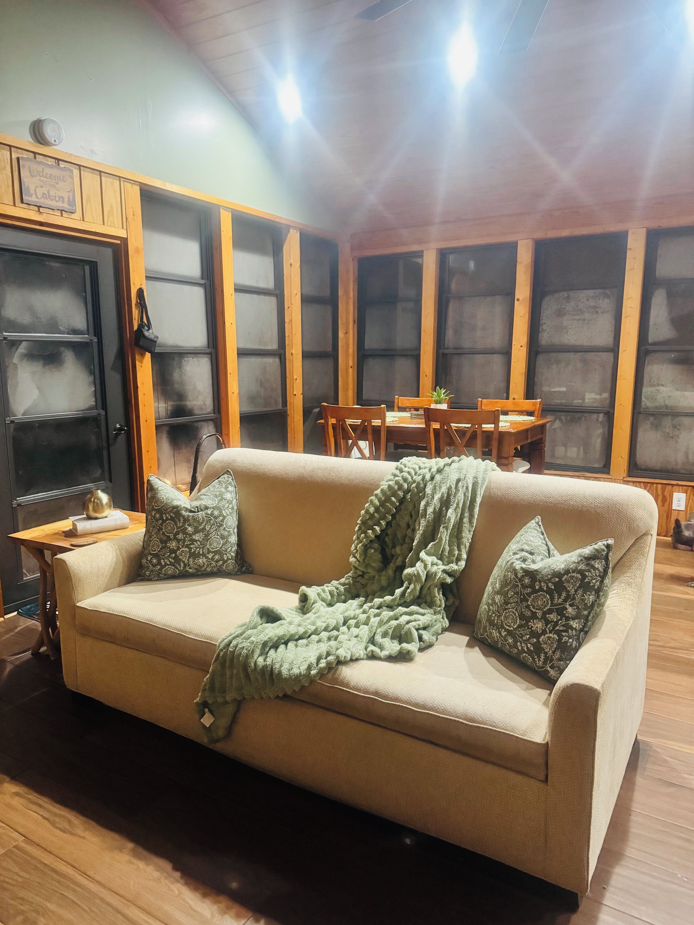 A cozy living room with a beige sofa, a green blanket, and patterned pillows. There is a wooden dining table with chairs, and a black door with a sign above it.