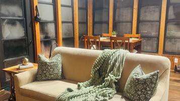 A cozy living room with a beige sofa, a green blanket, and patterned pillows. There is a wooden dining table with chairs, and a black door with a sign above it.