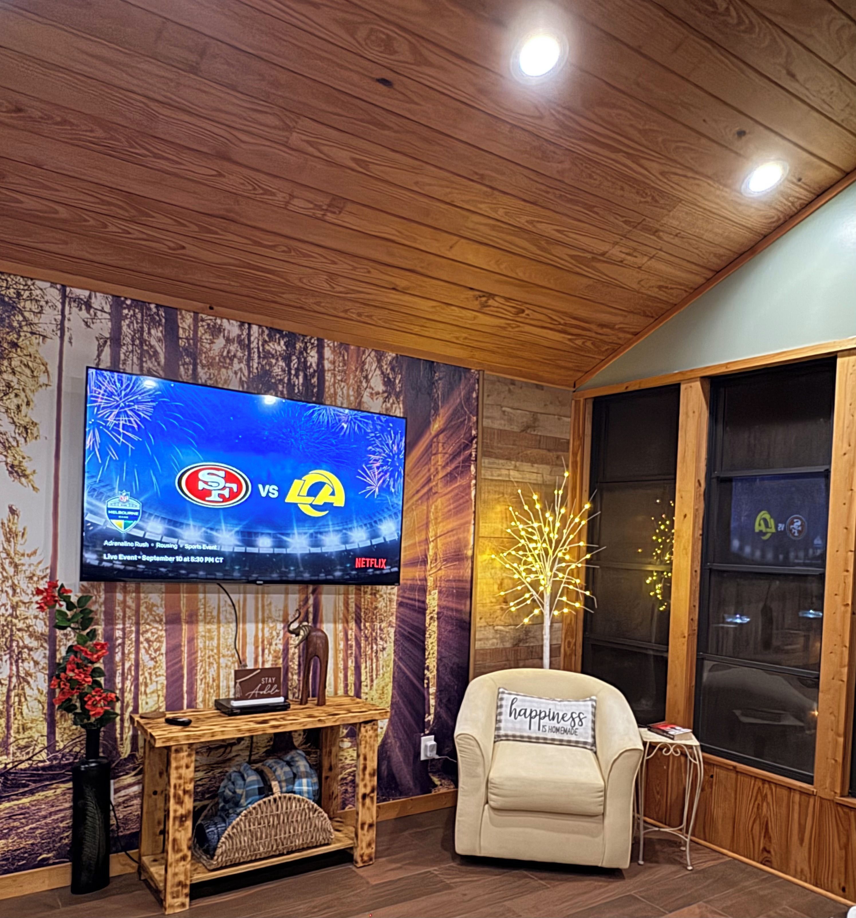 Ein Wohnzimmer mit einem an einer Holzwand befestigten Flachbildfernseher, einem bequemen Sessel, einem kleinen Holztisch mit einer Vase und einer Fernbedienung sowie einem dekorativen, beleuchteten Baum.