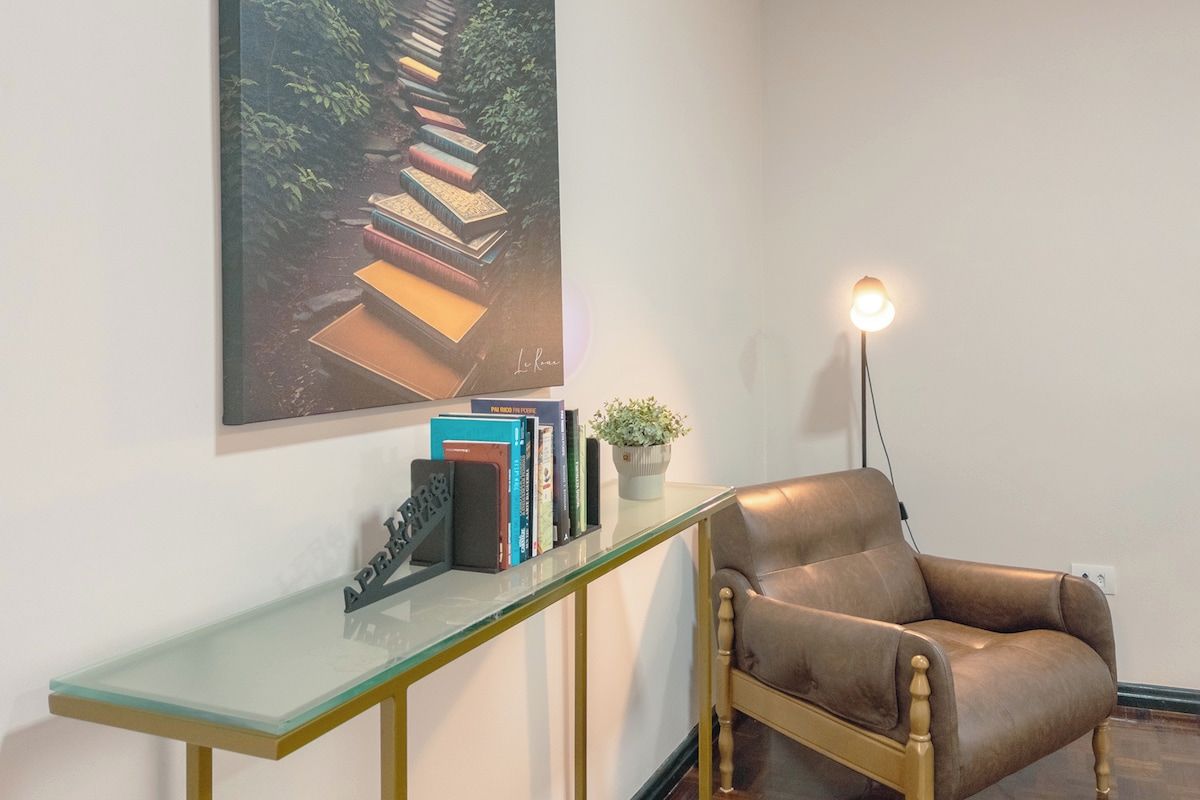 Quarto com mesa de centro em vidro, uma cadeira de couro marrom, luminária de parede e quadro emoldurado.