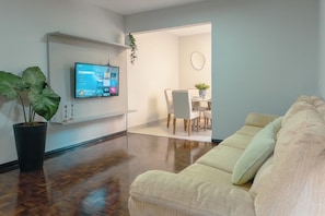 A living room with a flat-screen TV mounted on the wall, a large potted plant, a beige sofa, and a dining area with a table and chairs. - Comfort and Top Location in Maringa - MJ41 (Maringá)