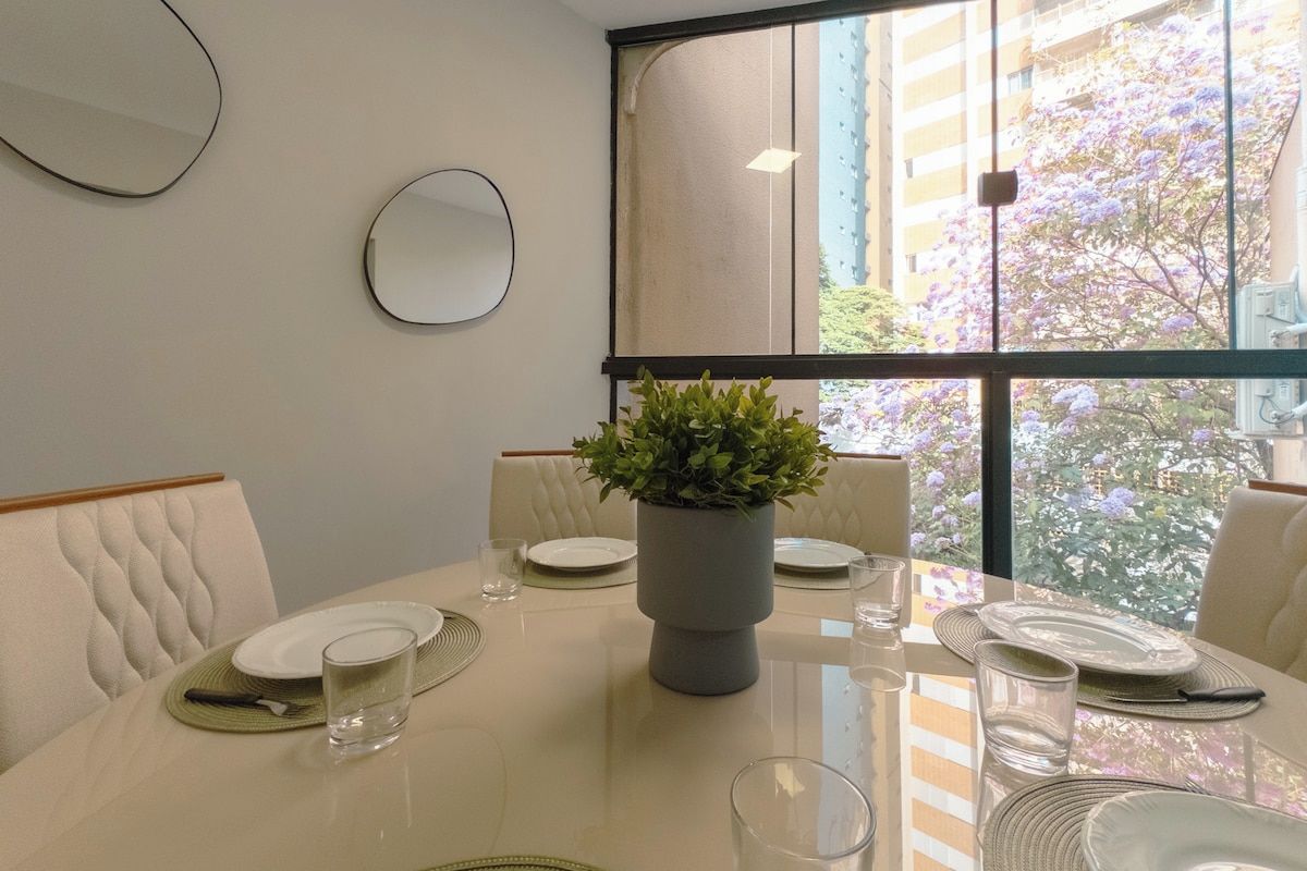 A dining table set for four with a potted plant in the center, white walls, and a view of buildings and trees outside.