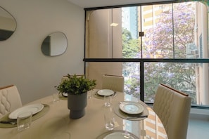 A dining area with a table set for four, a potted plant, and a view of buildings and trees through large windows. - Comfort and Top Location in Maringa - MJ41 (Maringá)