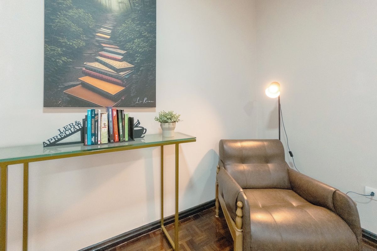 Una habitación con un sillón de cuero, una mesa de vidrio con libros, una planta pequeña y un cuadro de un sendero en el bosque.