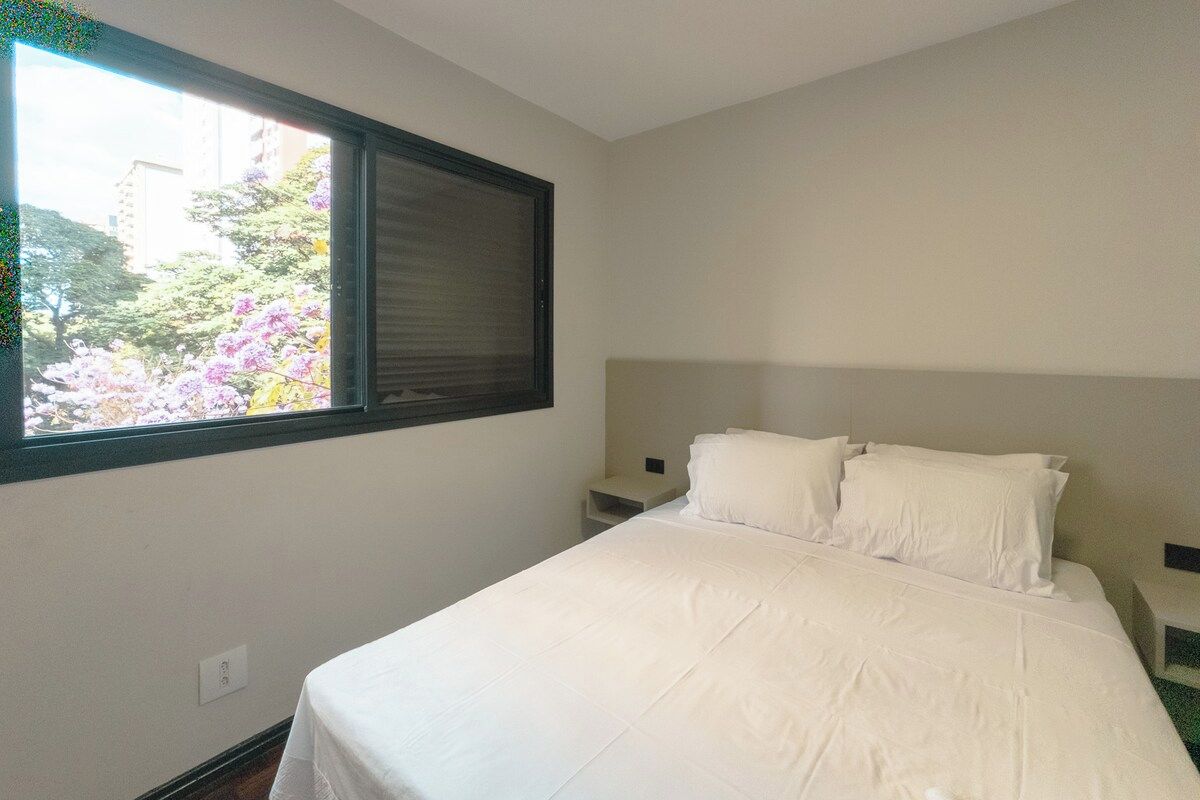 A hotel room with a single bed, a nightstand, a window with blinds, and a view of trees and buildings outside.