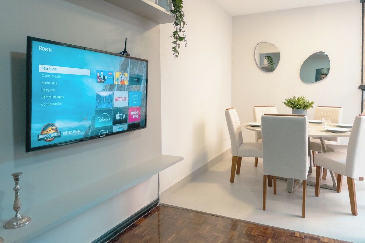 Uma sala de estar moderna com televisão de tela plana exibindo a interface Roku, uma área de jantar com cadeiras brancas e uma mesa, e decoração minimalista.