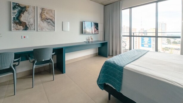 Habitació d'hotel amb un llit, una taula de treball, dues cadires, una finestra gran i obra d'art a la paret.