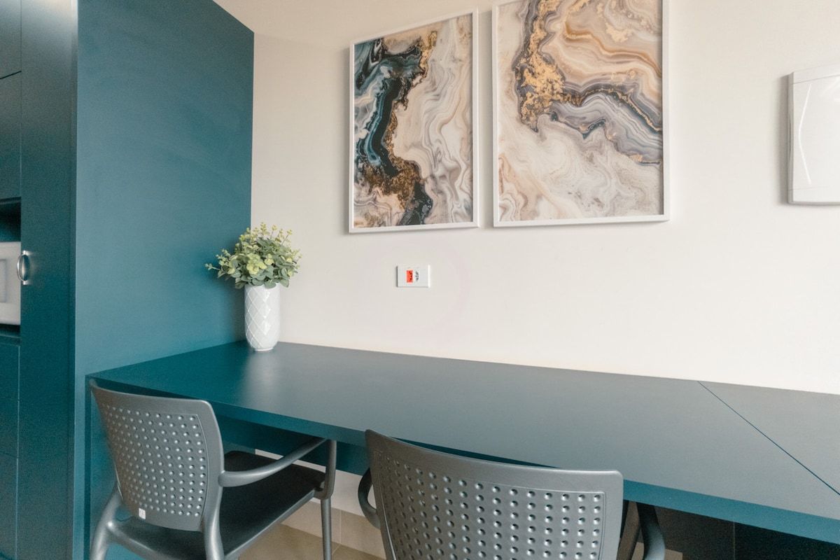 A modern dining area with a dark teal table and two perforated chairs. There are two abstract paintings on the wall above the table.