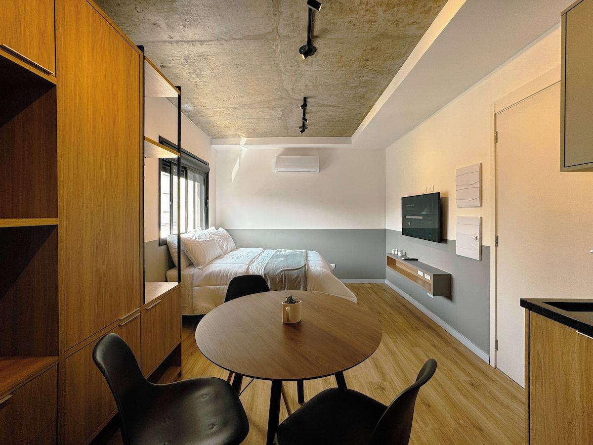 A modern hotel room with a bed, a round table with chairs, a wardrobe, and a television on the wall.