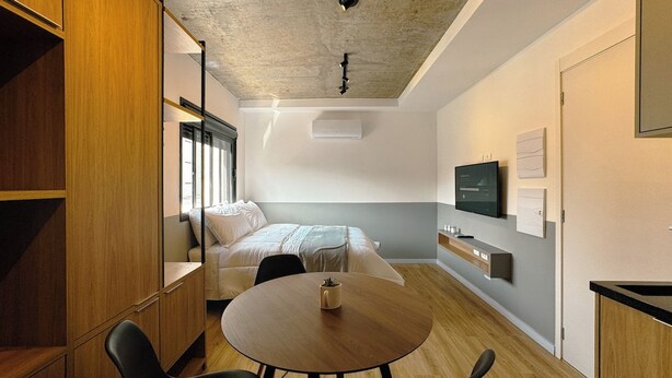 A modern hotel room with a bed, a round table with chairs, a wardrobe, and a television on the wall. - Venha conhecer este ape! - BE-DEO-403 (Maringá)