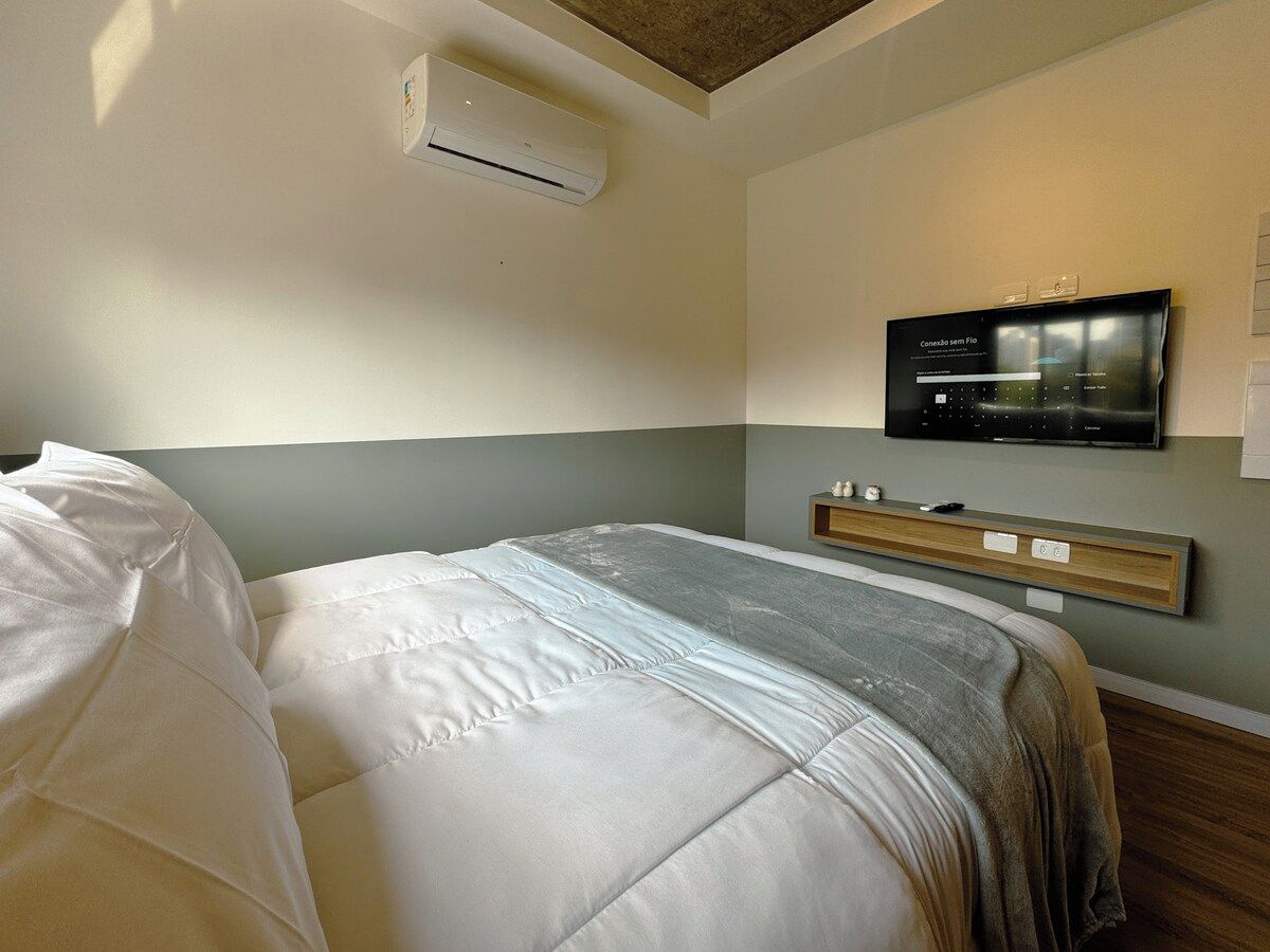 A hotel room with a bed, an air conditioning unit, a mounted television, and a bedside table.