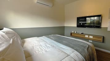 A hotel room with a bed, an air conditioning unit, a mounted television, and a bedside table.