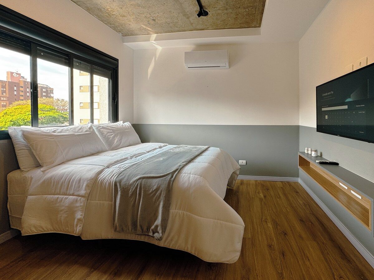 A modern bedroom with a bed, a television mounted on the wall, and a window providing a view of buildings and trees.