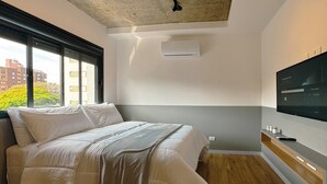 A modern bedroom with a bed, a television mounted on the wall, and a window providing a view of buildings and trees. - Venha conhecer este ape! - BE-DEO-403 (Maringá)