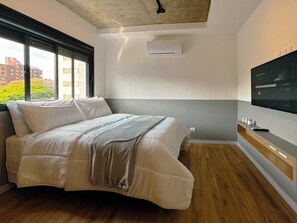 Ein modernes Schlafzimmer mit einem Bett, einem an der Wand montierten Fernseher und einem Fenster mit Blick auf Gebäude und Bäume.