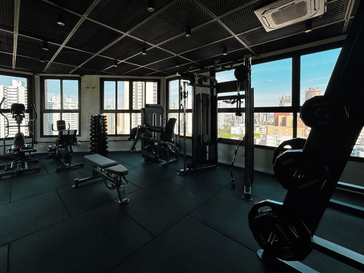 Ein Fitnessstudio mit unterschiedlichen Trainingsgeräten, großen Fenstern und Blick auf die Stadtlandschaft.