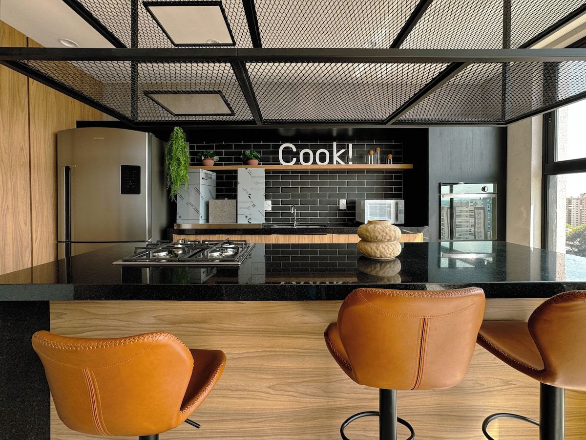 Una cocina moderna con encimera negra, electrodomésticos de acero inoxidable y un cartel que dice 'Cook!'. Hay tres sillas marrones en primer plano.