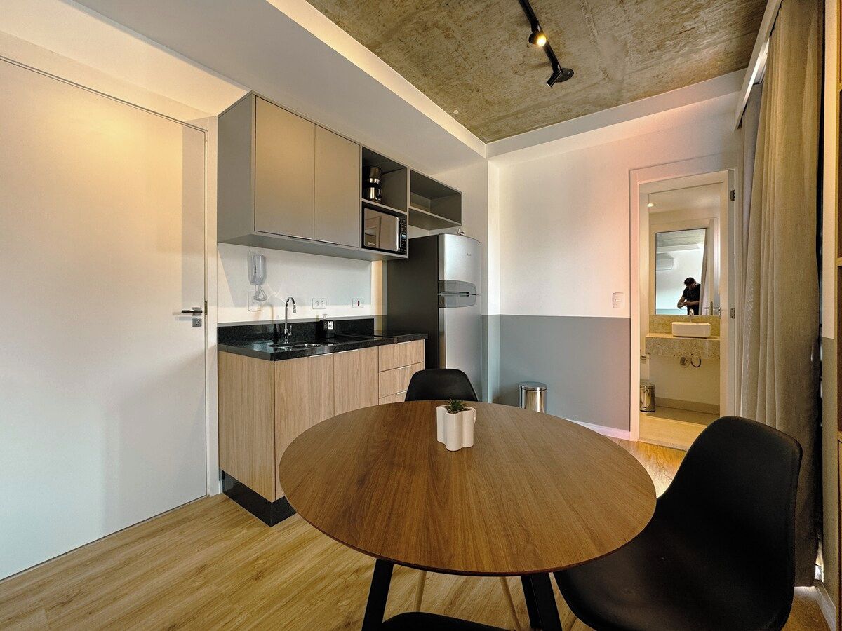 A compact kitchen with modern appliances, a dining area with a round table, and a visible bathroom in the background.