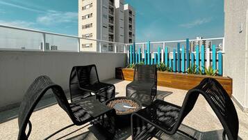 A rooftop terrace with wicker furniture and a fire pit, overlooking a cityscape.