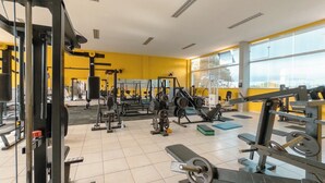 Academia com diversos equipamentos de ginástica, amplas janelas e uma parede amarela.