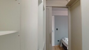 Corredor estreito com uma cama no final, um guarda-roupa e uma porta que dá para outro quarto.