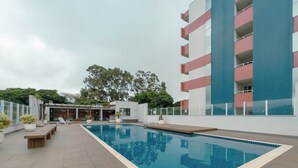 Área de piscina com espreguiçadeiras, bancos e um edifício com varandas.