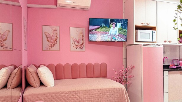 Quarto em tons de rosa com cama, televisão e espaço de cozinha.