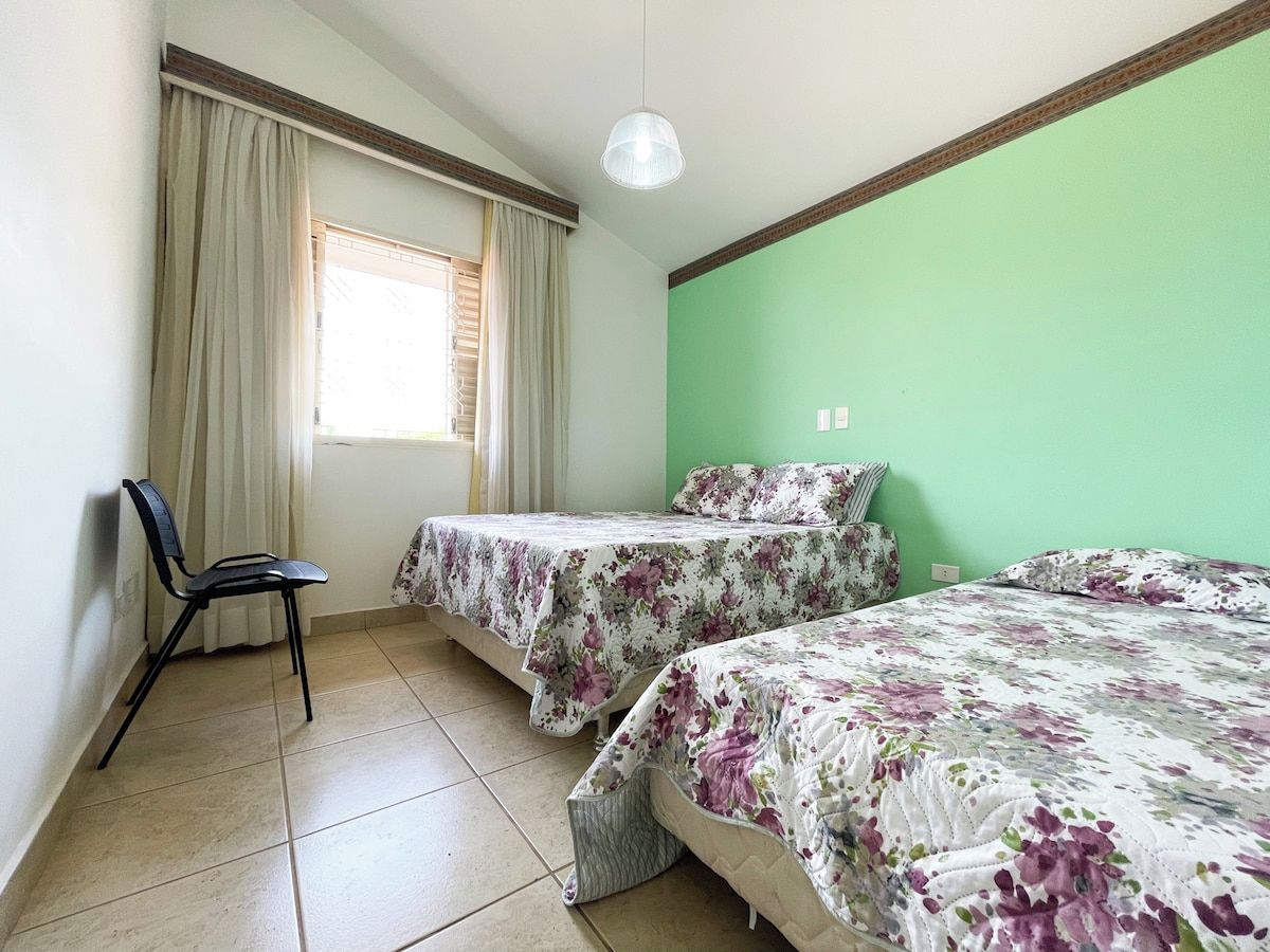 Quarto com duas camas, uma cadeira, janela com cortinas e parede verde.