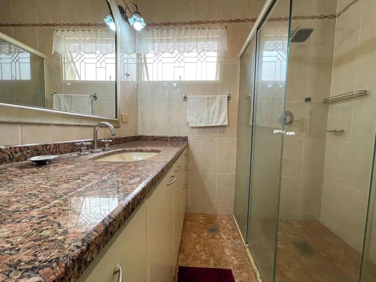 Casa de banho com lavatório bancada em granito, cabine de duche em vidro e janela com cortinas de renda.