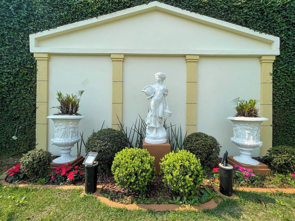 Um jardim com uma estátua, dois vasos com plantas e um edifício branco com colunas.