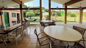 Um pátio externo com mesa de vidro, cadeiras de vime e vista para um jardim.