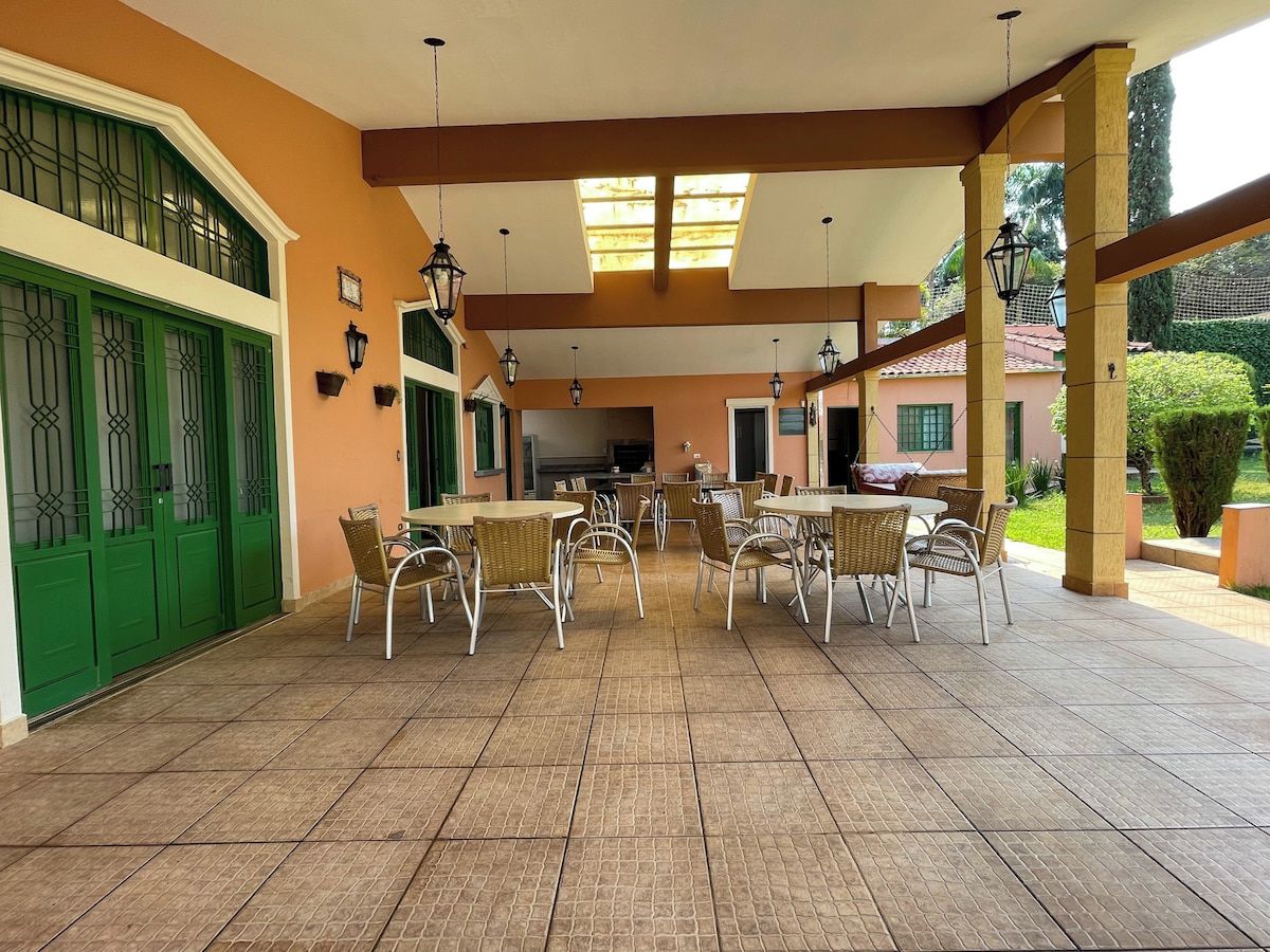 Um alpendre coberto com várias mesas e cadeiras, piso em azulejos e uma porta verde.