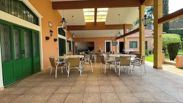 Um alpendre coberto com várias mesas e cadeiras, piso em azulejos e uma porta verde.