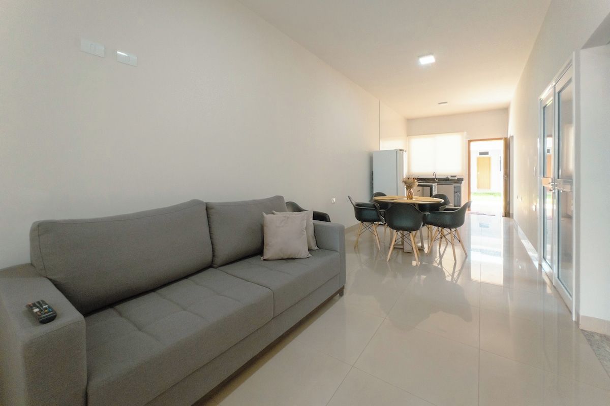 Uma sala de estar moderna com um sofá cinzento, uma área de jantar com mesa e cadeiras, e uma cozinha ao fundo.