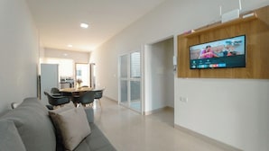 Uma sala de estar moderna com um sofá cinza, uma TV de tela plana fixada em uma parede de madeira e um espaço de jantar com mesa e cadeiras.
