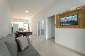 A modern living room with a grey sofa, a flat-screen TV mounted on a wooden wall, and a dining area with a table and chairs. - Imperial House with 3 Bedrooms in Maringa - BON705 (Maringá)