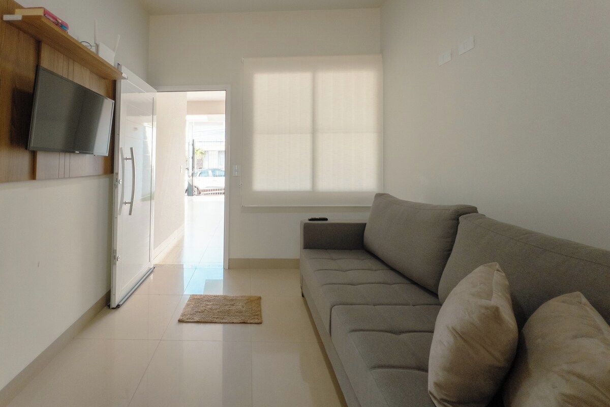 A living room with a sofa, a flat-screen TV mounted on a wooden panel, a window with blinds, and a door leading to another room.